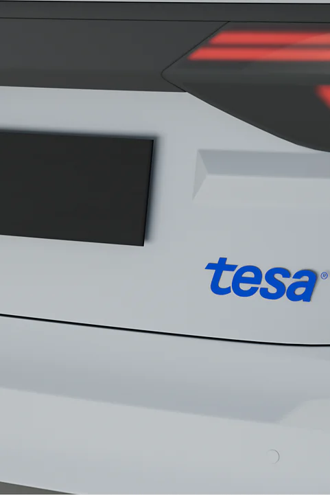 The image shows the back of a white vehicle. The license plate is covered. A small round camera is visible above it. The logo tesa in blue letters is displayed on the right side of the vehicles rear. (This text has been generated by AI)