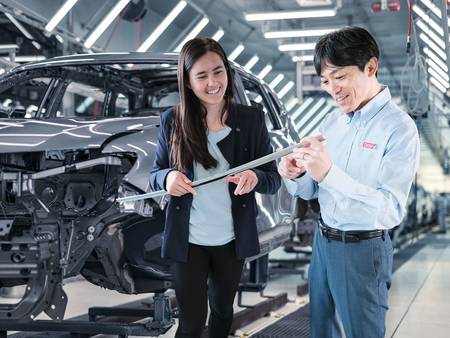 Two individuals stand in an automotive factory. One holds a sheet of tesa tape, and they both smile while discussing. Behind them, an unfinished car chassis is visible. Bright overhead lights illuminate the scene. (This text has been generated by AI)