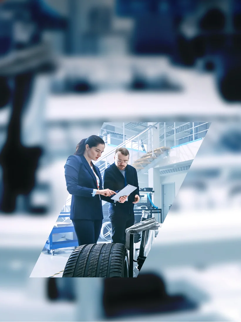 Two individuals review documents in an automotive workshop. A car chassis with exposed wheels is visible in the foreground, with strips of tesa tape securing various components. The background is a modern, well-lit industrial space filled with equipment and tools. (This text has been generated by AI)